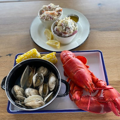 A whole steamed lobster, corn, and a pot of steamed clams on a tray next to a plate with slaw, melted butter, and potato salad