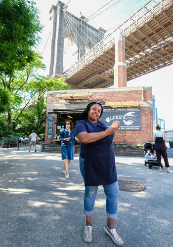 A smiling woman holding a lobster roll poses in front of the Brooklyn Bridge Luke's Lobster Shack